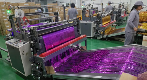 Our workers are operating advanced fringe cutting machines. They closely monitor the equipment, adjust parameters, and allow the metallic foil film to be rapidly and evenly cut into fine, dense tinsel strips. Long fringes continuously emerge from the machine as workers check the output in real time to ensure cutting precision and high production efficiency.